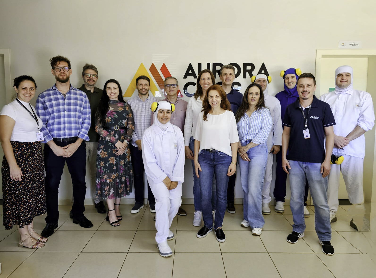 Foto posada com 15 pessoas aparecendo. Quatro estão usando roupas brancas e protetores auriculares. Na parede ao fundo aparece o logo da Cooperativa Aurora.