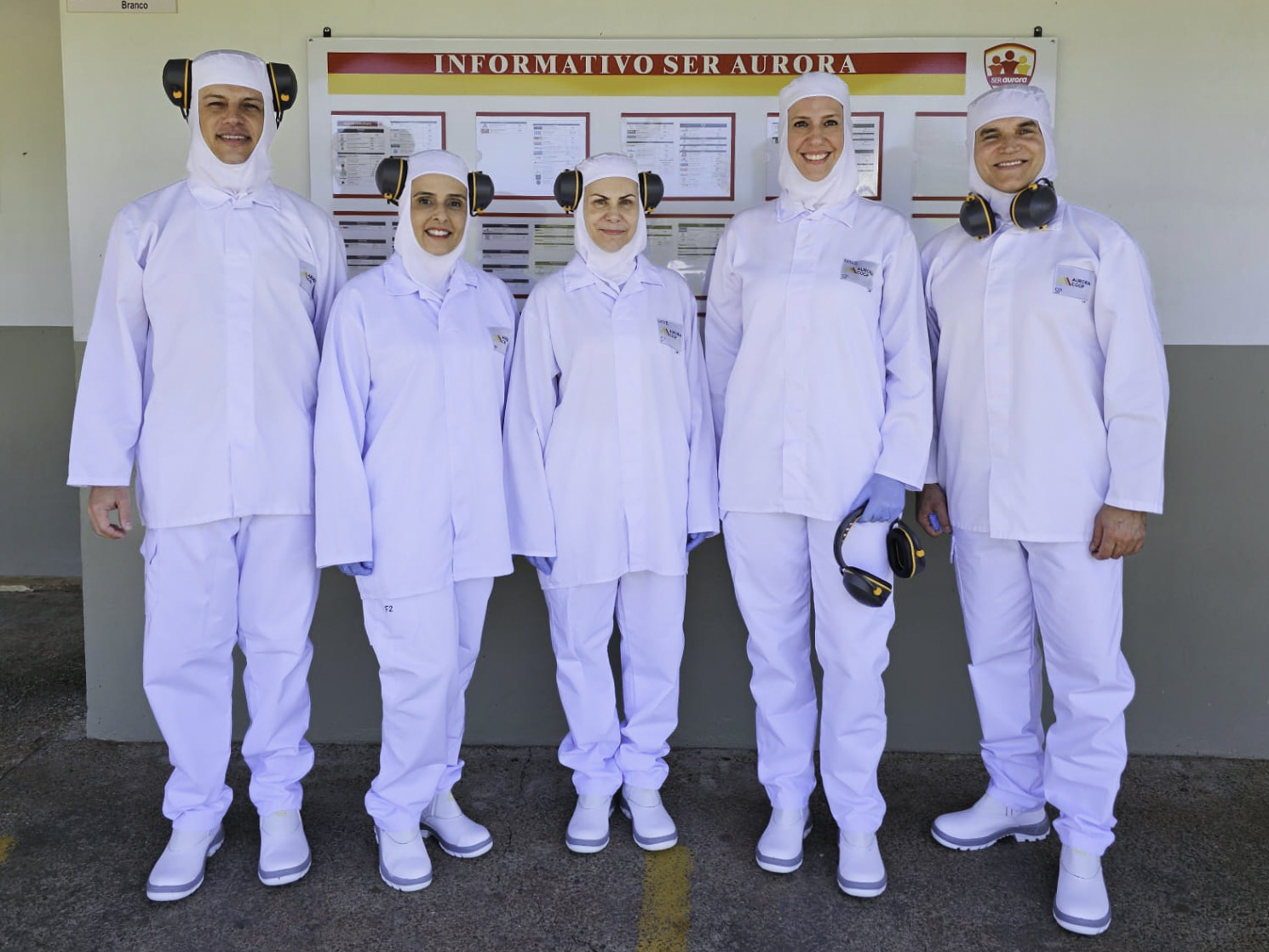Foto posada de cinco pessoas usando roupas brancas de proteção individual e protetores auriculares. Na parede ao fundo aparece o letreiro "informativo aurora" e um quadro de avisos.