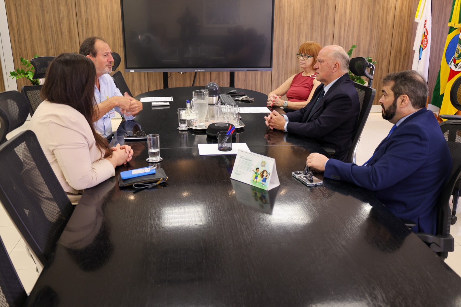 A imagem mostra a reunião formal, com os cinco participantes sentados  ao redor de uma mesa. O ambiente possui bandeiras institucionais e um monitor desligado ao fundo. Claudir gesticula enquanto fala e os demais escutam atentamente.