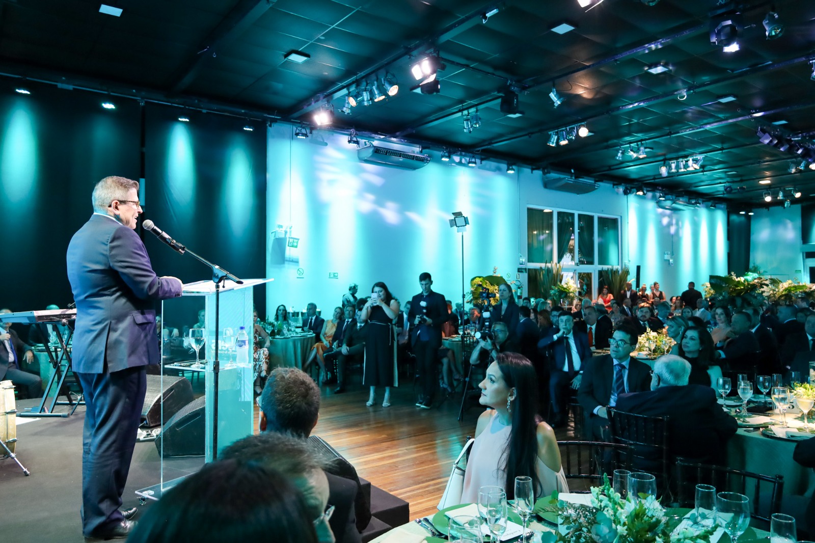 A foto mostra o novo presidente do Simers discursando. Ele aparece meio de costas, num púlpito, com o público de fundo.