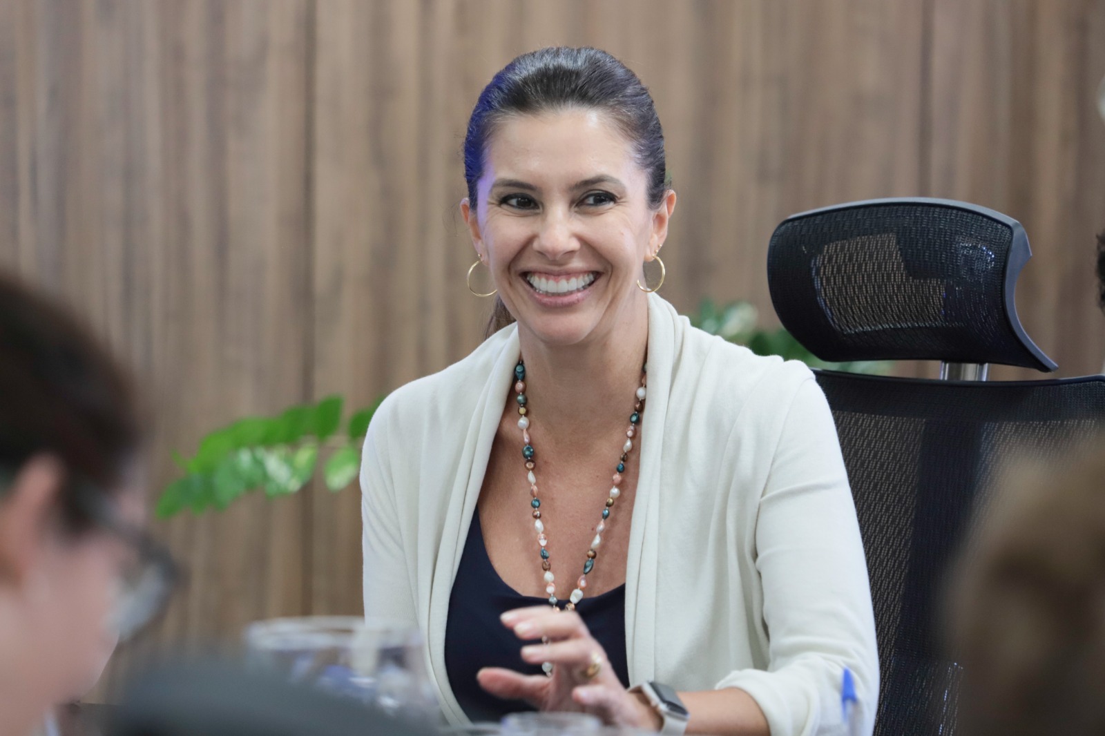 Aline Fagundes é branca, tem cabelo preto liso. Está sorrindo e gesticula. Usa blusa preta e casaco branco.