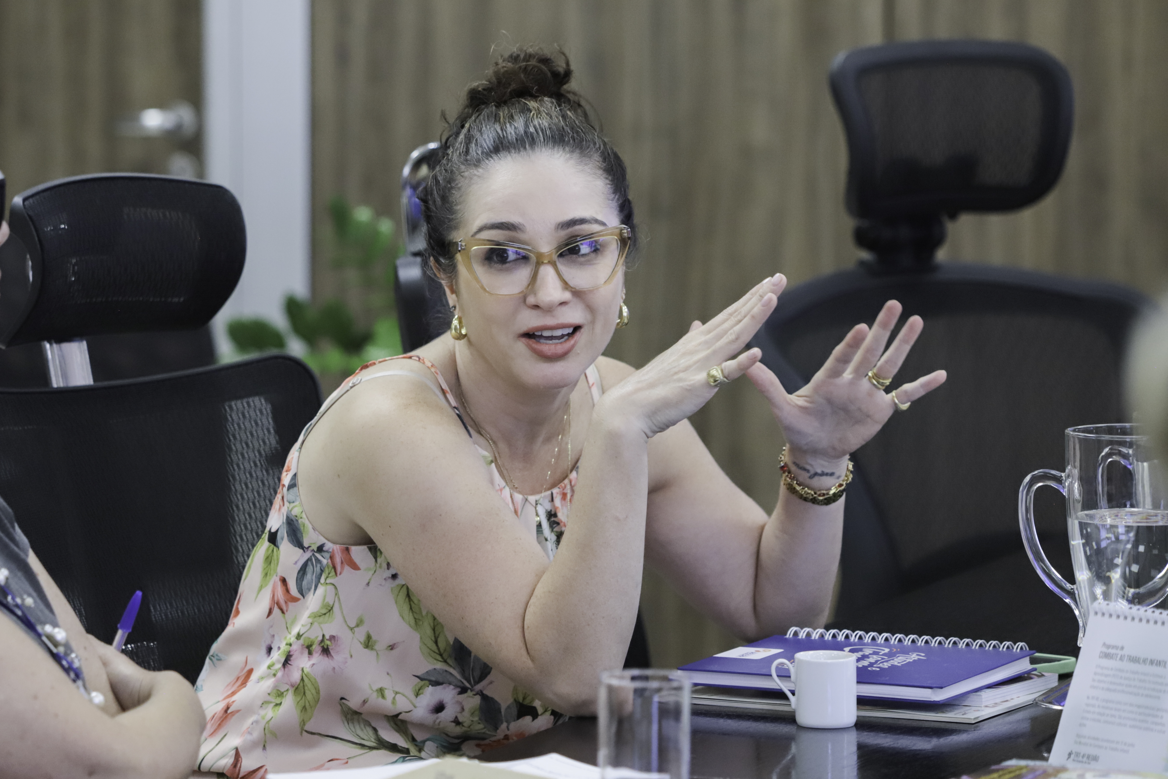 Thais Bruch é branca, tem cabelo preto liso, usa vestido floreado e óculos de armação laranja;