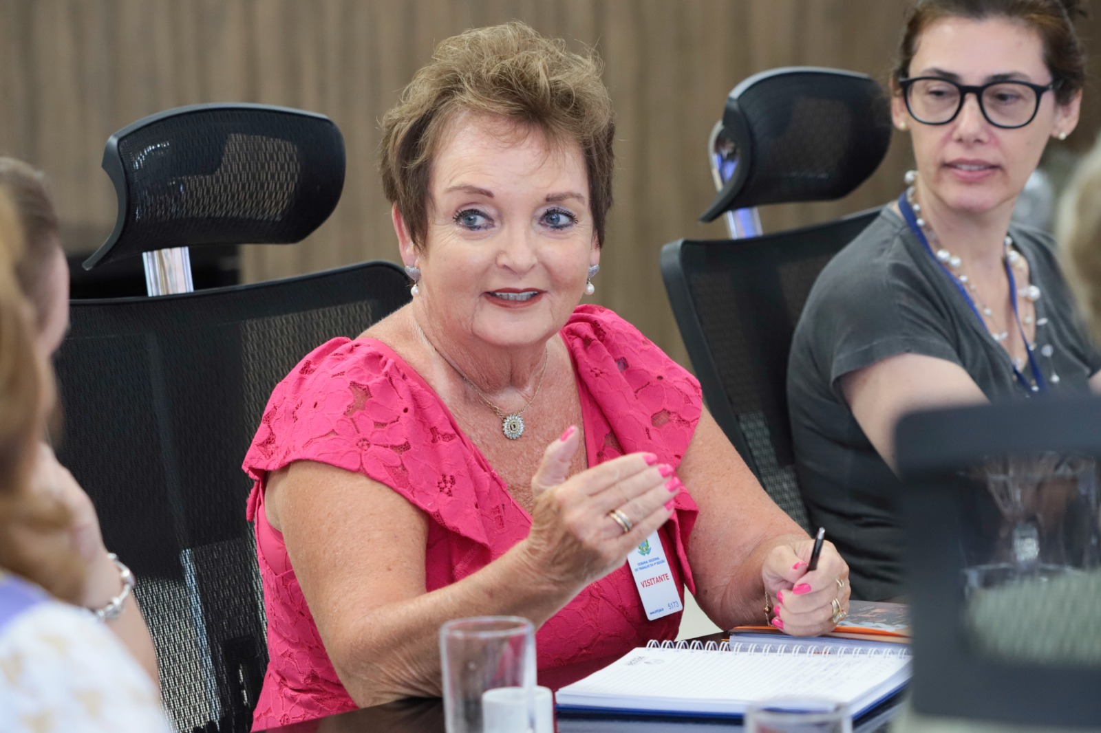 Denise Gonzalez é branca, tem cabelo castanho curto, olhos claros, usa blusa rosa e está gesticulando.