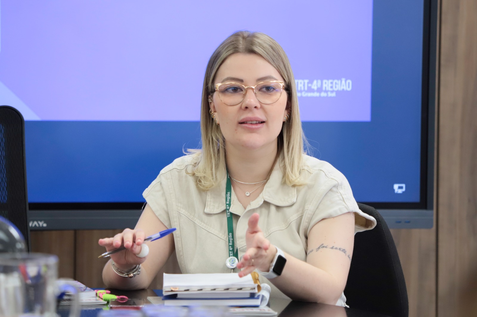 Lais Gerhardt é branca, loira, usa óculos de armação amarela. Está gesticulando. Ao fundo aparece uma tela ligada escrito "TRT-4ª Região"