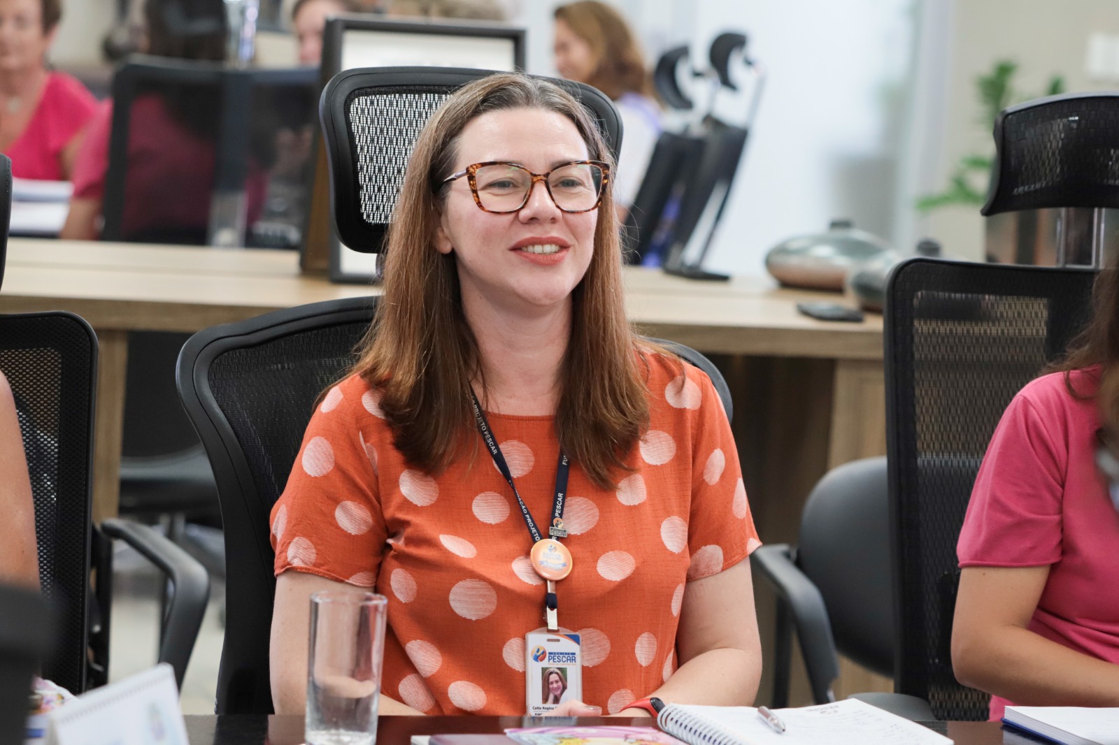 Cátia Beulke é branca, tem cabelo ruivo e usa blusa laranja com círculos brancos. usa óculos com armação laranja e preta. Fala e está sorrindo.