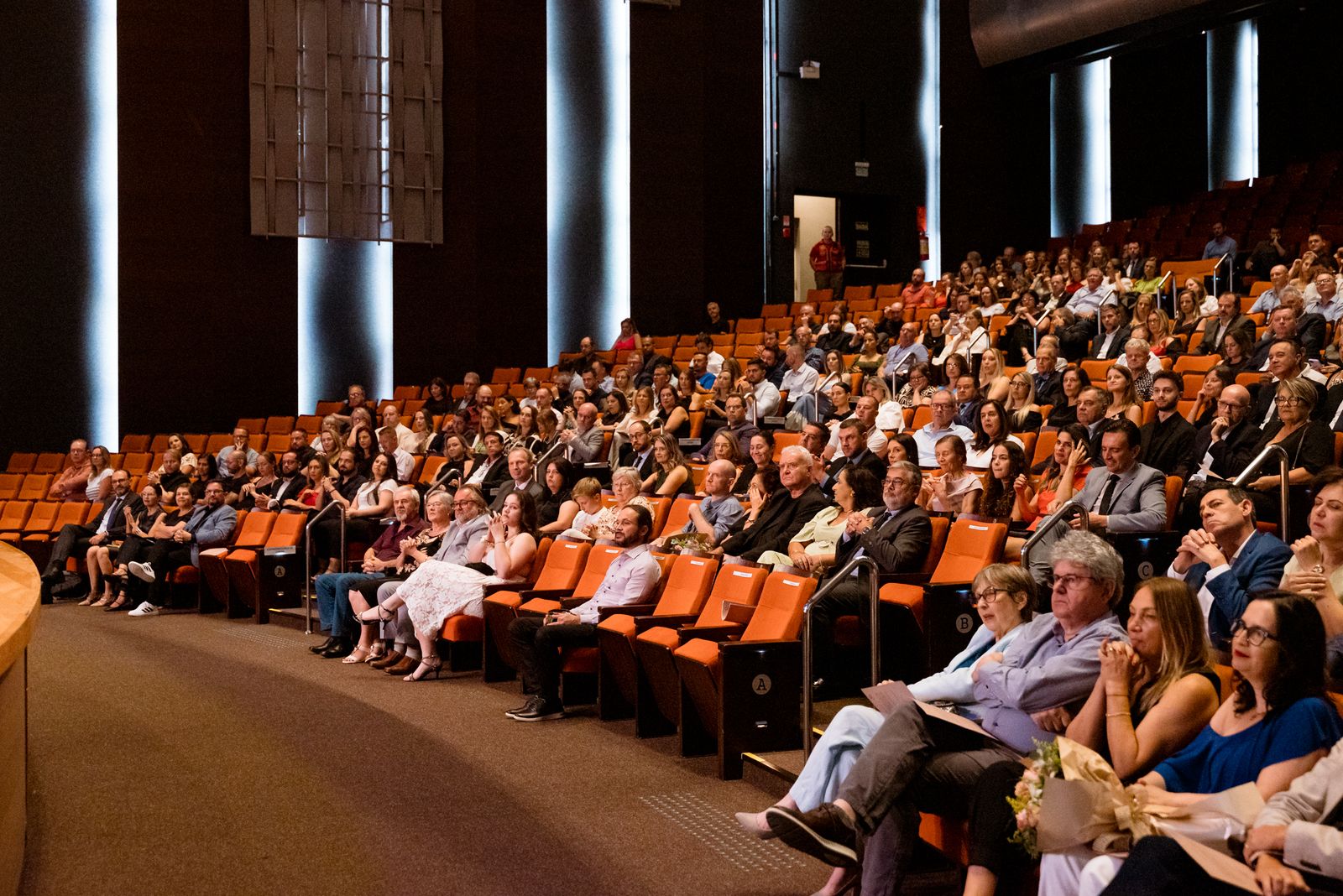 Teatro da Univates lotado, a plateia sentada em bancos laranja presta atenção.f