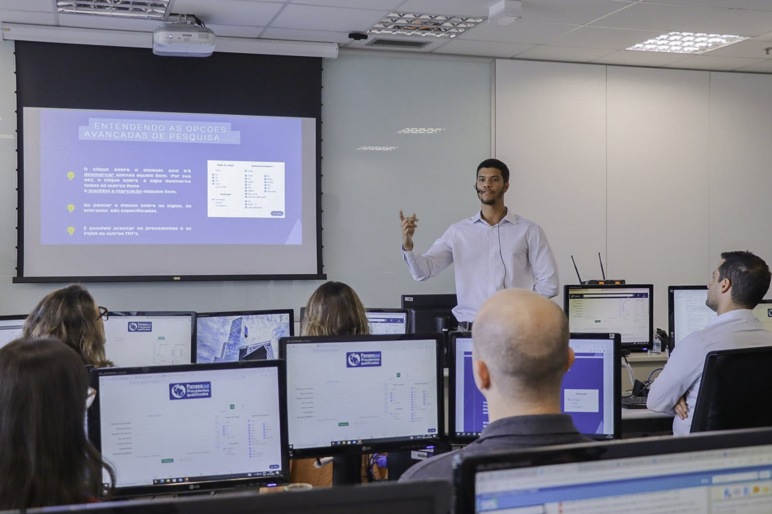 Foto de uma sala de aula com várias pessoas de costas em primeiro plano, usando computadores. Ao fundo, Marcelo Souza está de pé falando e gesticulando ao lado de uma tela de projeção.