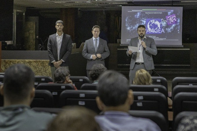 Público aparece de costas em primeiro plano, ao fundo três pessoas estão de pé falando. Atrás deles há uma tela com o logo do Pangea