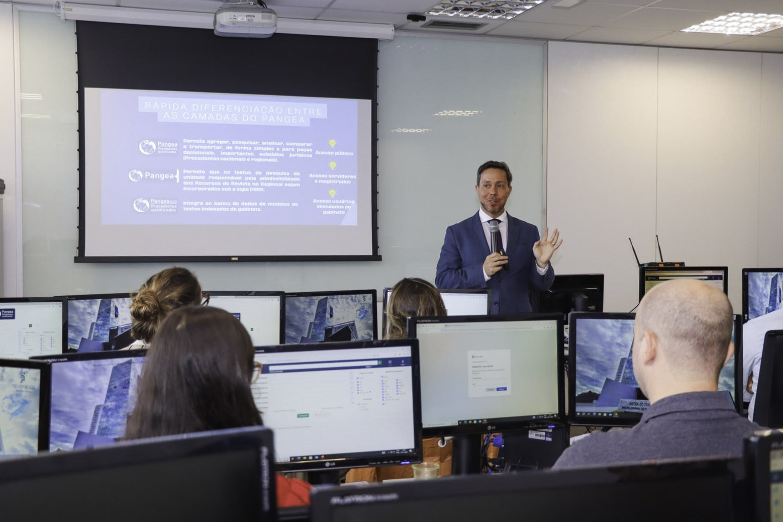 Foto de uma sala de aula com várias pessoas de costas em primeiro plano, usando computadores. Ao fundo, Rodrigo Trindade está de pé falando e gesticulando ao lado de uma tela de projeção.