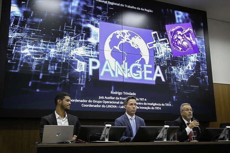 Três pessoas aparecem na mesa oficial de um evento. Ao fundo, aparece um grande tela com o logo do Pangea.
