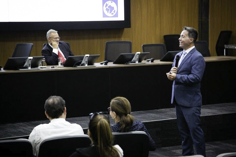 Rodrigo Trindade está de pé, na direita da imagem, falando e sorrindo. Na esquerda aparecem pessoas no público e ao fundo, na mesa oficial, uma pessoa está sorrindo.