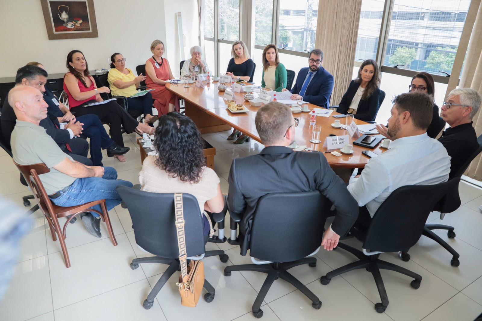 Foto dos participantes na mesa de negociação por outro ângulo. Ao fundo aparece uma quadro na parede e no lado direito está a janela. Em primeiro plano aparece alguém falando, de costas, e os demais estão olhando para ele.