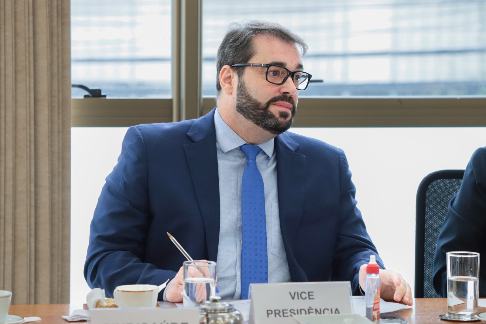 Alexandre Corrêa da Cruz é branco, usa óculos de aro escuro, cabelo e barba pretos. Ele usa um blazer azul marinho e camisa e gravata azuis em tons mais claros. À sua frente há uma placa onde está escrito "Vice-Presidência".