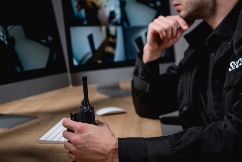 Vigilante em frente a monitores e segurando um walkie-talkie. Ele usa uniforme preto.