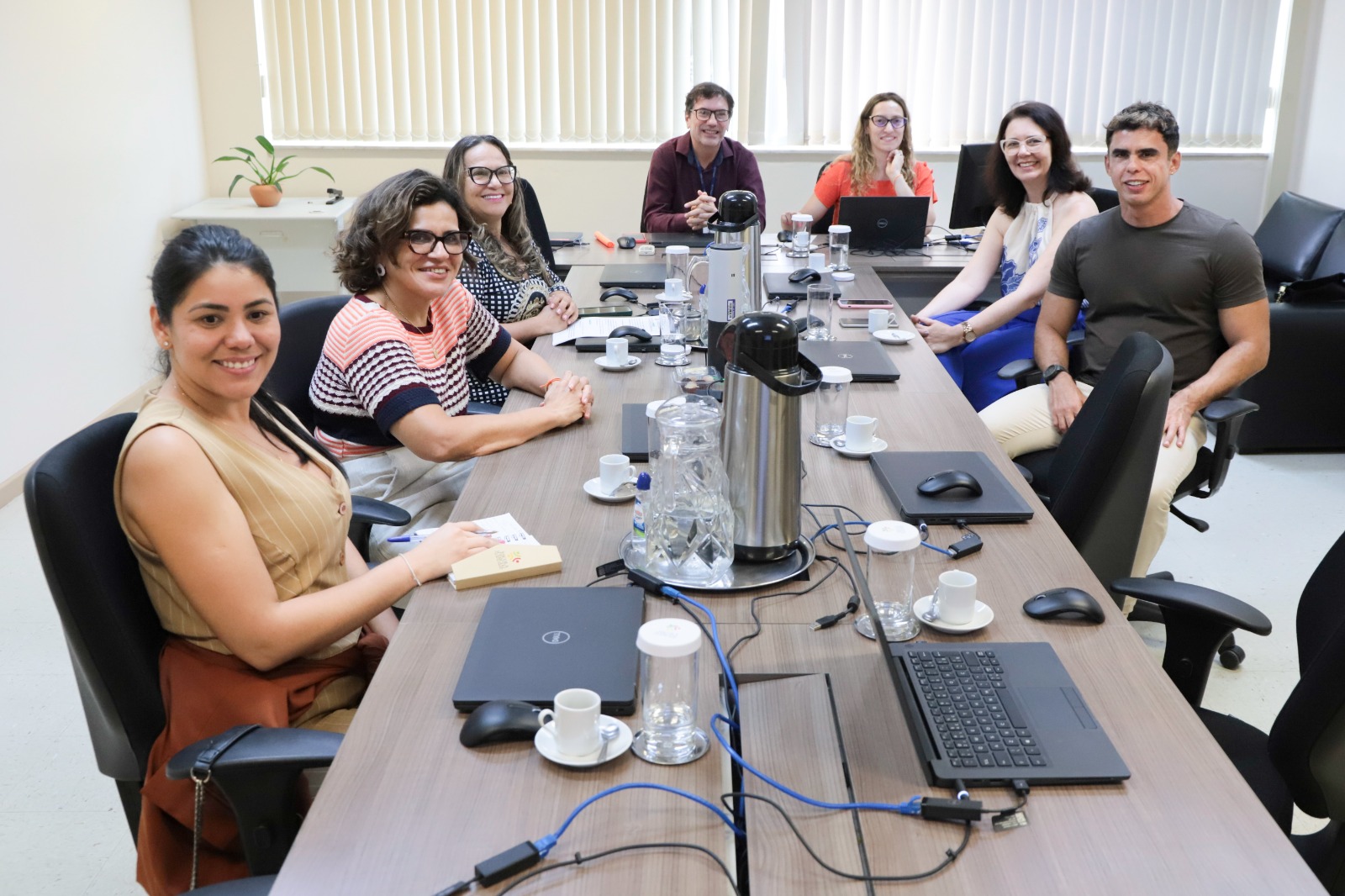 Sete pessoas aprecem sentadas em torno de uma mesa retangular. Três do lado esquerdo, dois ao fundo, e dois do lado direito. Todos olham em direção à câmera e sorriem.