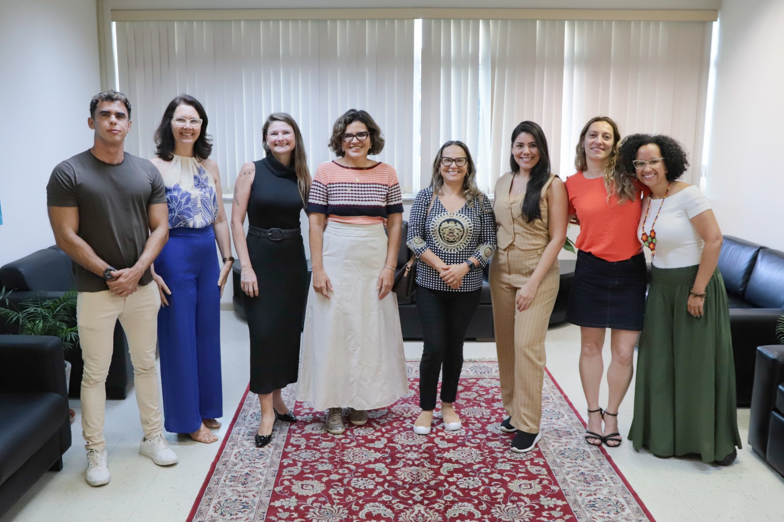 Oito pessoas aparecem de pé, posando para a foto, sorrindo. Elas estão sobre um tapete decorativo. Nas laterais aparecem sofás e ao fundo uma janela com as cortinas fechadas.
