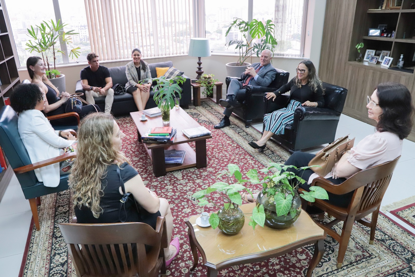Oito pessoas aparecem de pé, posando para a foto, sorrindo. Elas estão sobre um tapete decorativo. Nas laterais aparecem sofás e ao fundo uma janela com as cortinas fechadas.