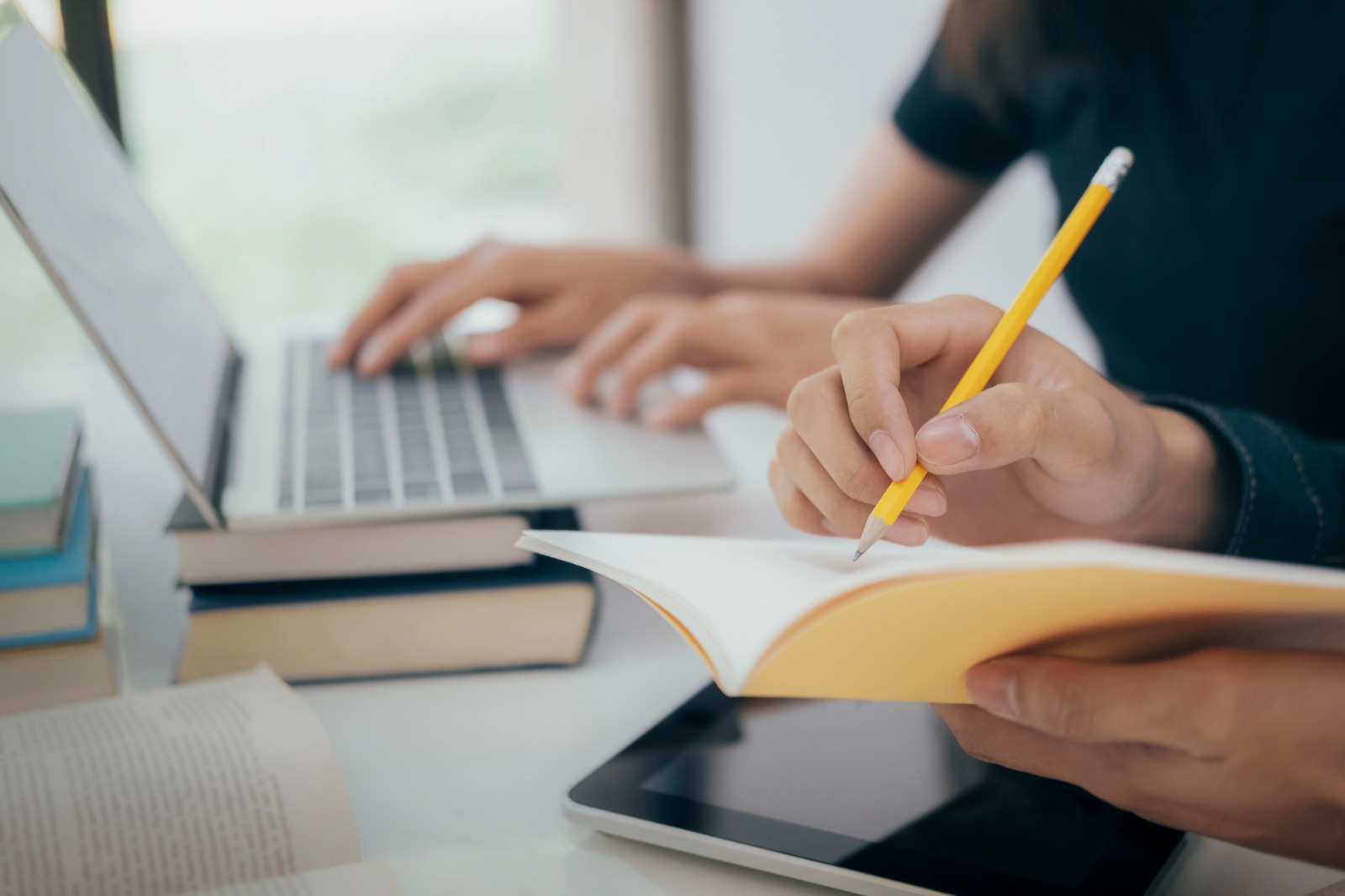 Foto ilustrativa de uma pessoa usando um notebook e outra fazendo anotações em um caderno com um lápis.