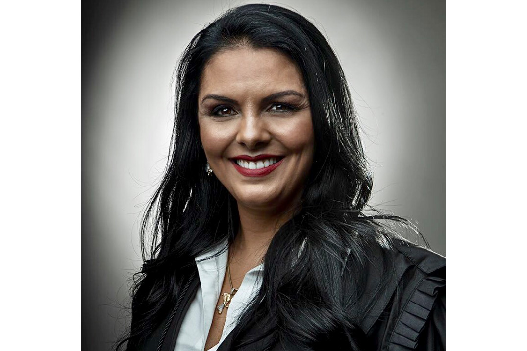 Tânia Reckziegel é parda, tem cabelo preto liso e está sorrindo para a foto. Usa camisa branca e casaco preto.
