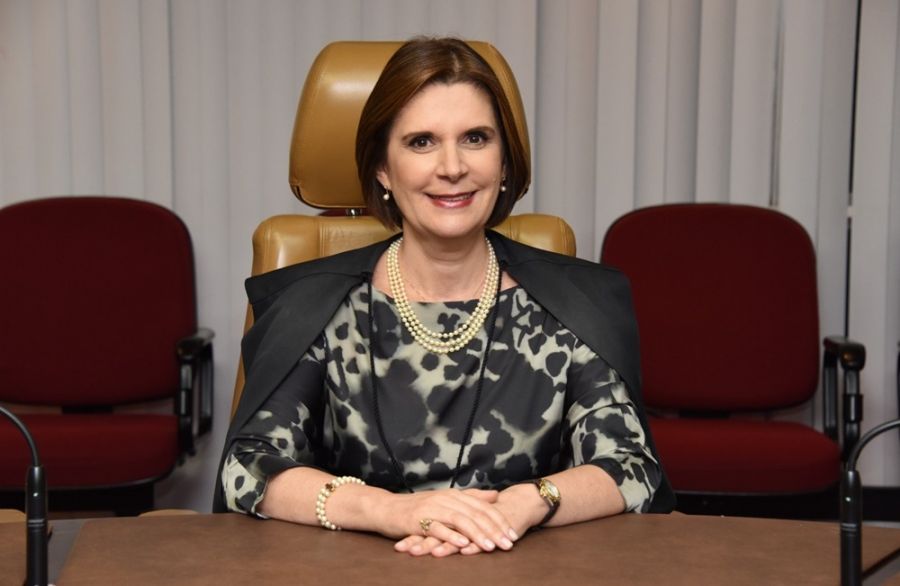 Ministra Maria Elizabeth Rocha é banca, tem cabelo curto castanho e usa vestido com estampas em verde e preto. Está sorrindo para a foto, com os braços sobre a mesa.
