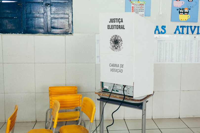 Urna eleitoral encoberta por proteção de papelão branco, com a inscrição “Cabine de votação” e brasão da Justiça Eleitoral.