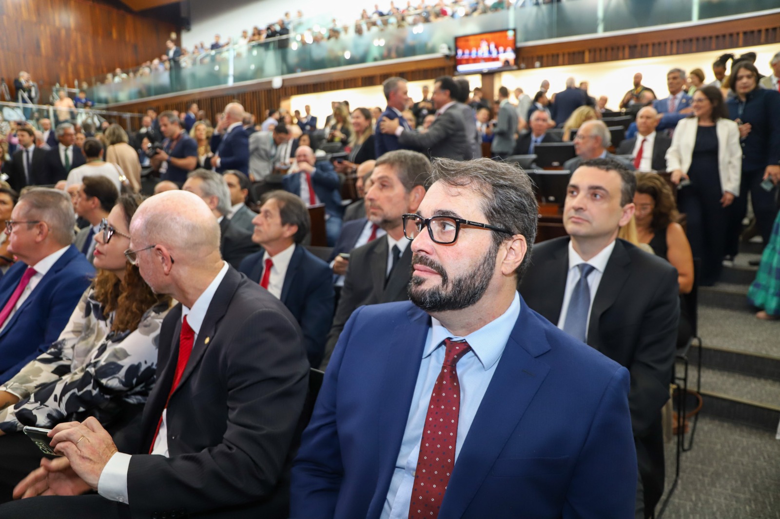 A imagem mostra o desembargador Alexandre em primeiro plano acompanhando a sessão solene da ALRS. A maioria dos participantes está vestida formalmente, com ternos e gravatas. O ambiente é um plenário com várias fileiras de assentos e uma galeria superior onde mais pessoas estão observando.