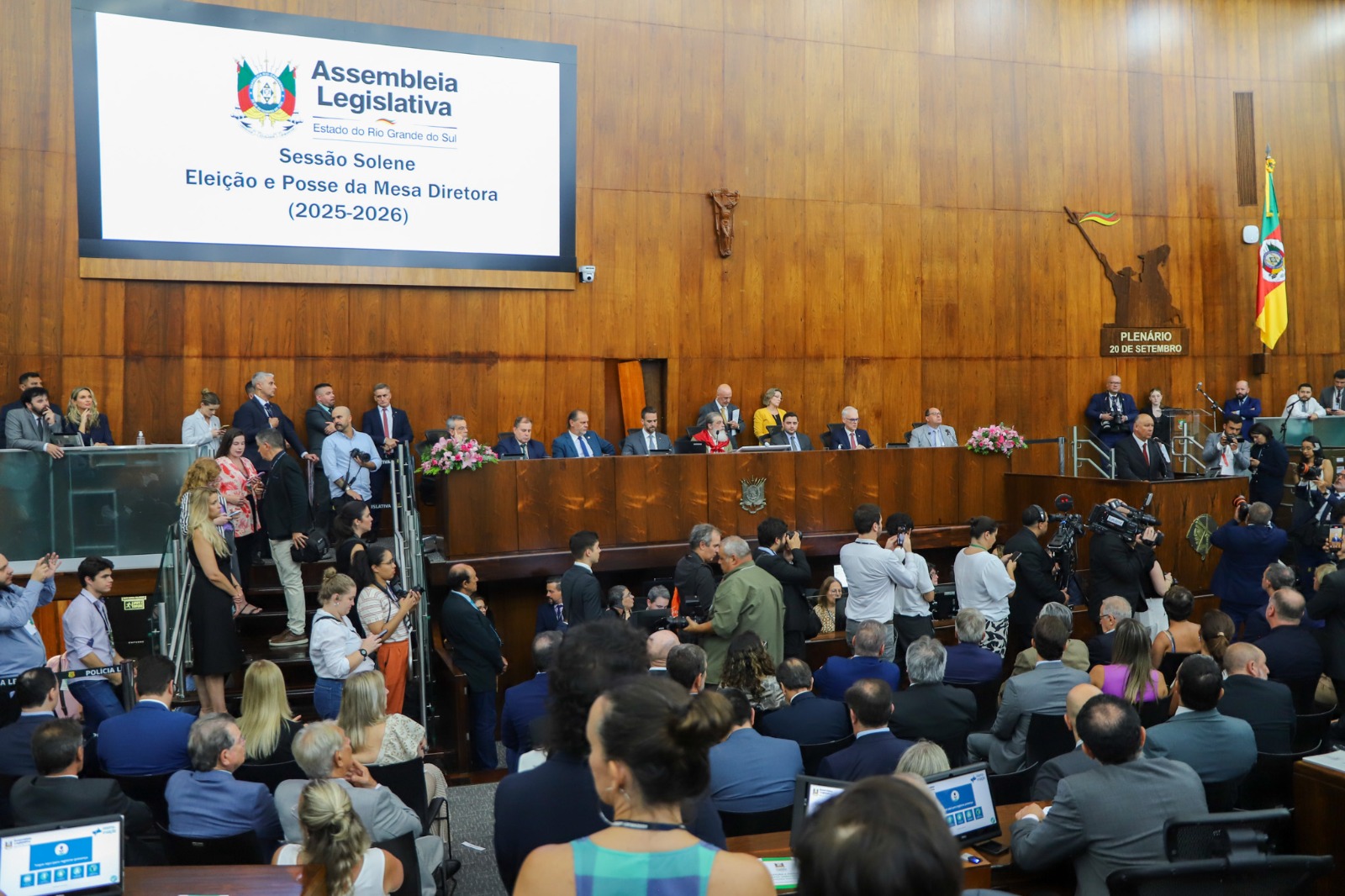 A imagem mostra o Plenário 20 de Setembro, da A, durante a cerimônia solene de eleição e posse da nova mesa diretora para o biênio 2025-2026. No destaque, autoridades e parlamentares estão posicionados na mesa diretora, enquanto o público ocupa as galerias e o plenário, com fotógrafos e cinegrafistas registrando o evento. O painel eletrônico exibe informações sobre a solenidade, e a decoração é marcada por arranjos de flores.