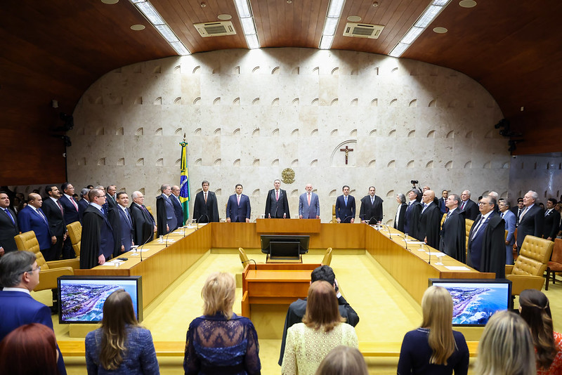A imagem mostra o plenário do Supremo Tribunal Federal (STF) durante uma cerimônia oficial. Ao centro, destaca-se a mesa principal com ministros e autoridades em posição de respeito, provavelmente durante a execução do Hino Nacional ou um momento solene. O ambiente é caracterizado pelo design moderno, com paredes em tom claro, o crucifixo e o brasão da República ao fundo, além da bandeira do Brasil. O público está posicionado em frente aos ministros, com algumas pessoas em pé e outras sentadas.
