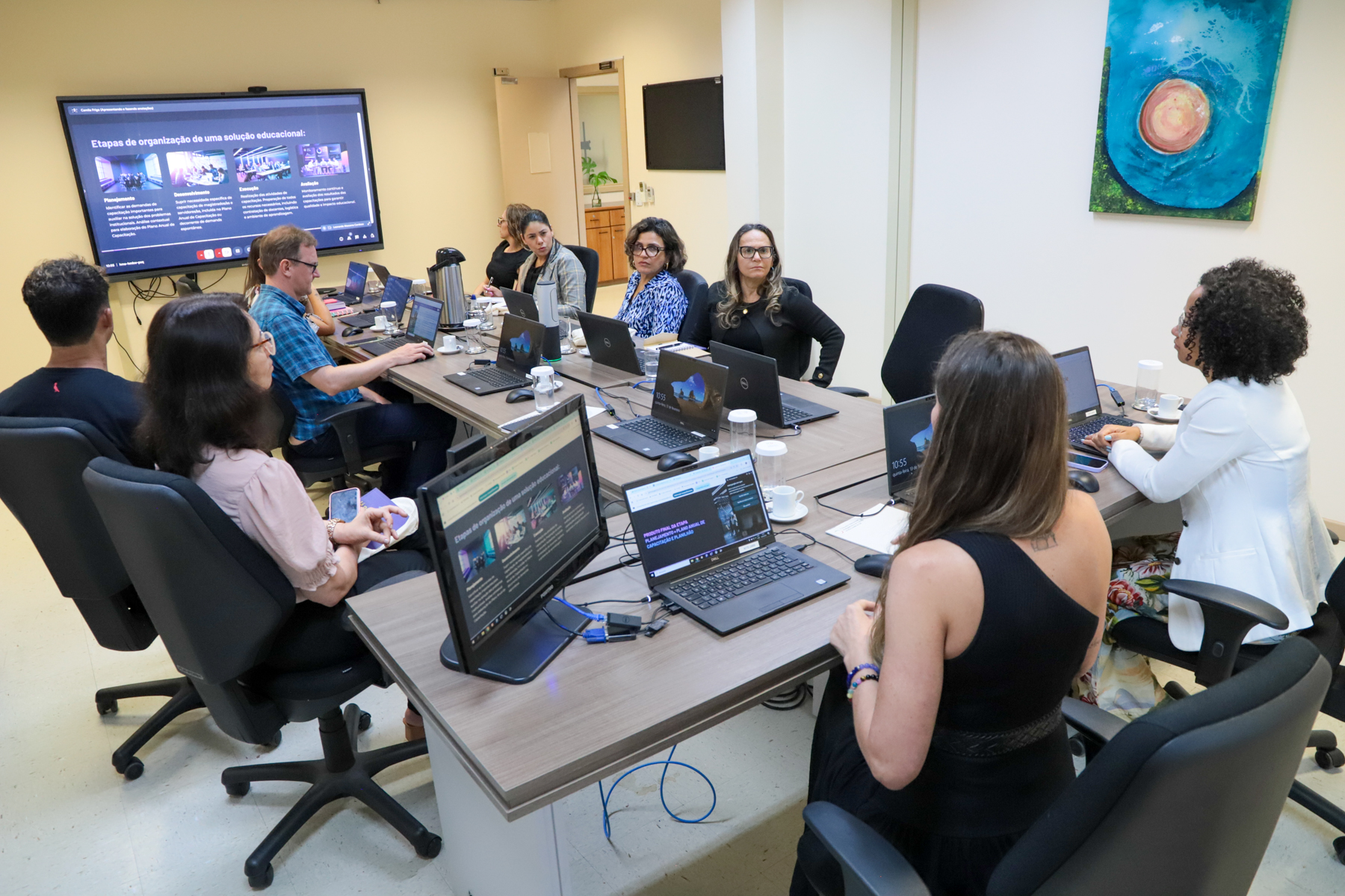 Reunião na sala do Conselho Consultivo, com os visitantes e servidores da EJud sentados à mesa. A secretária da EJud-4, Camila Frigo, conduz a reunião.