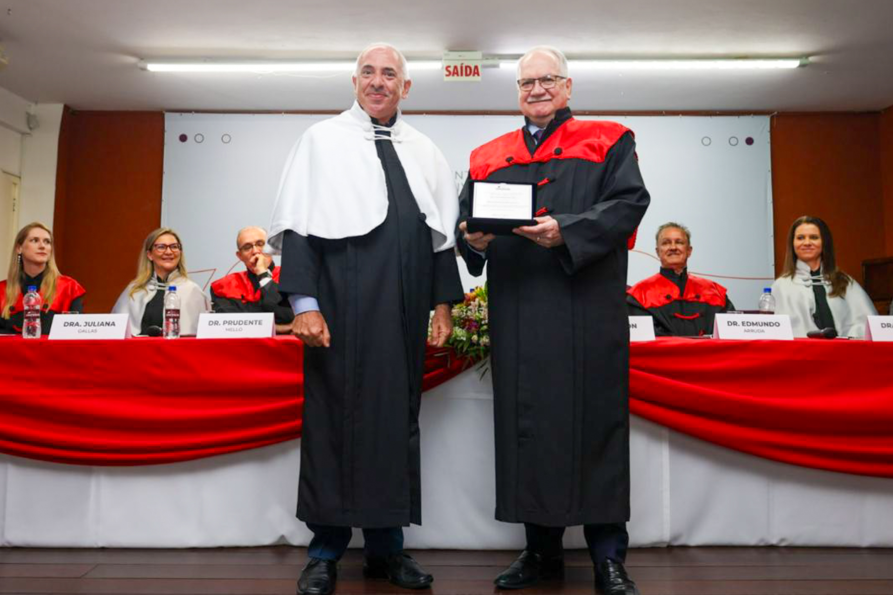 Ministro Fachin recebendo a outorga do título de “Professor Honoris Causa” no auditório do pelo Centro Universitário Cesusc (UNICESUSC). As cores predominantes são preto, vermelho e branco.