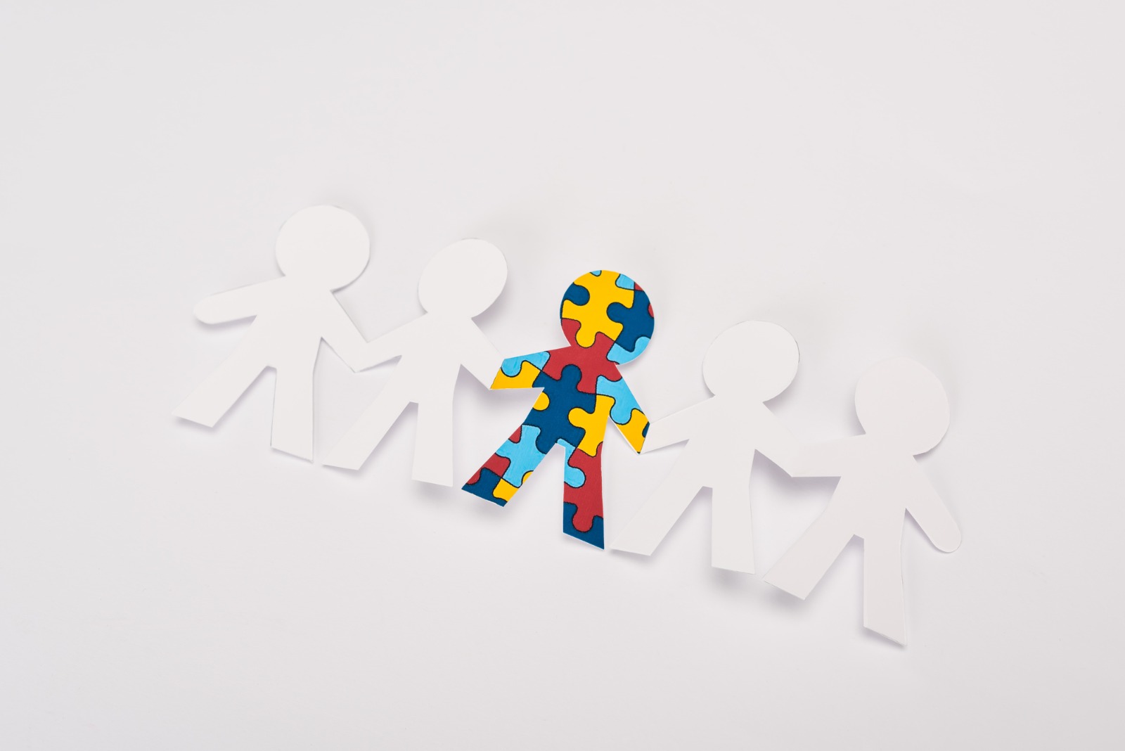 Imagem ilustrativa de cinco bonecos de papel de mãos dadas, sendo que o do meio está colorido com peças de quebra-cabeça, símbolo do autismo.