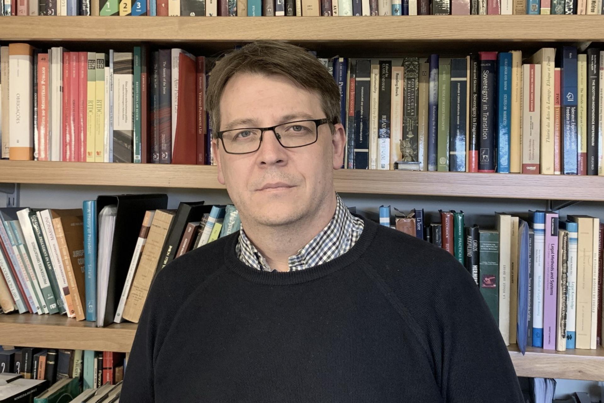 Professor Cláudio Michelon é um homem de meia idade. Tem a pele clara e cabelos curtos e castanhos. Ele usa suéter preto, sobre uma camisa xadrez branca e preta. Está em frente a uma estante com livros. Usa óculos de grau.