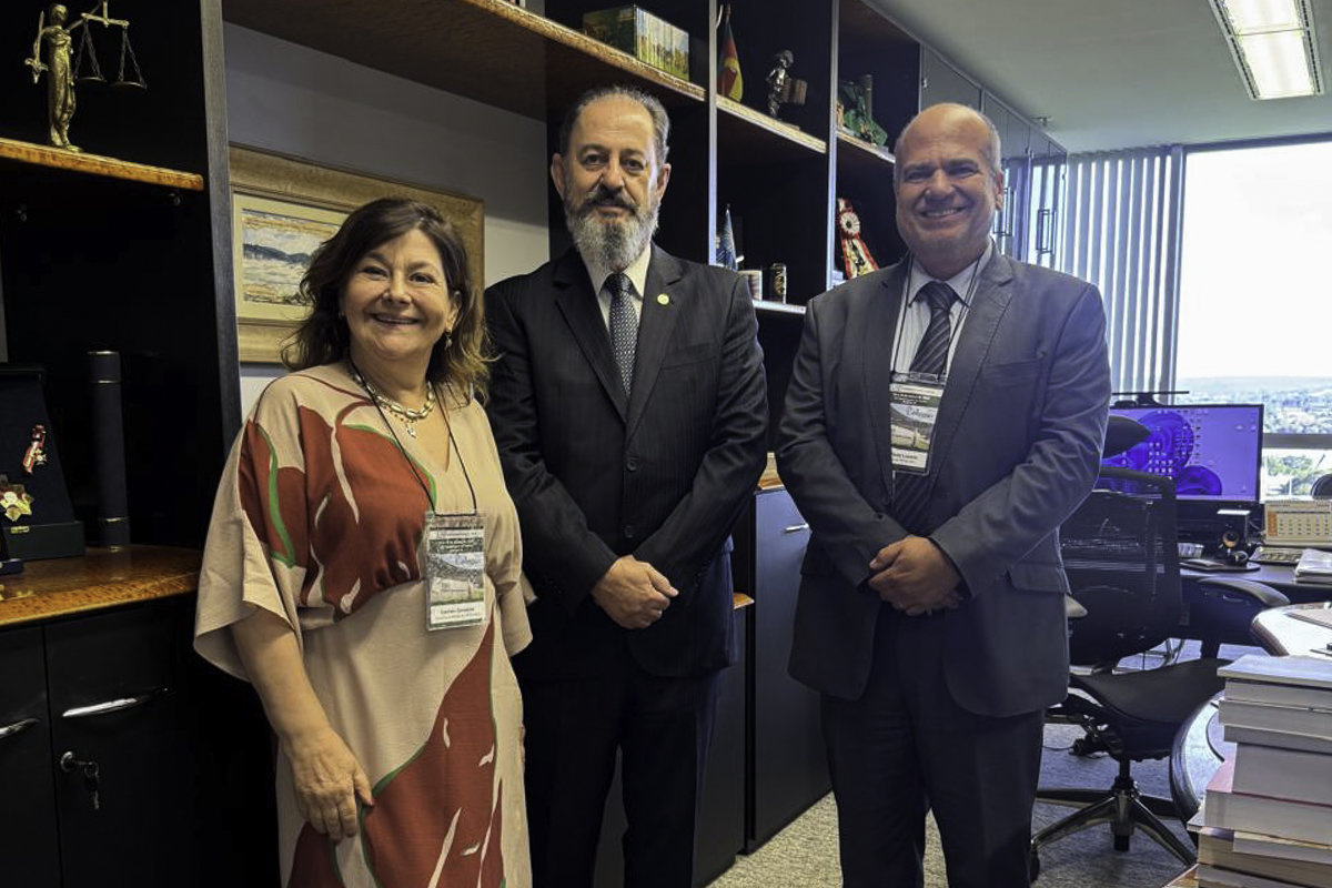 Carmen, Hugo e João Paulo posam sorrindo para a foto no gabinete do ministro. Ao fundo aparece uma estante com livros e objetos decorativos, a mesa de trabalho do ministro e uma janela. 