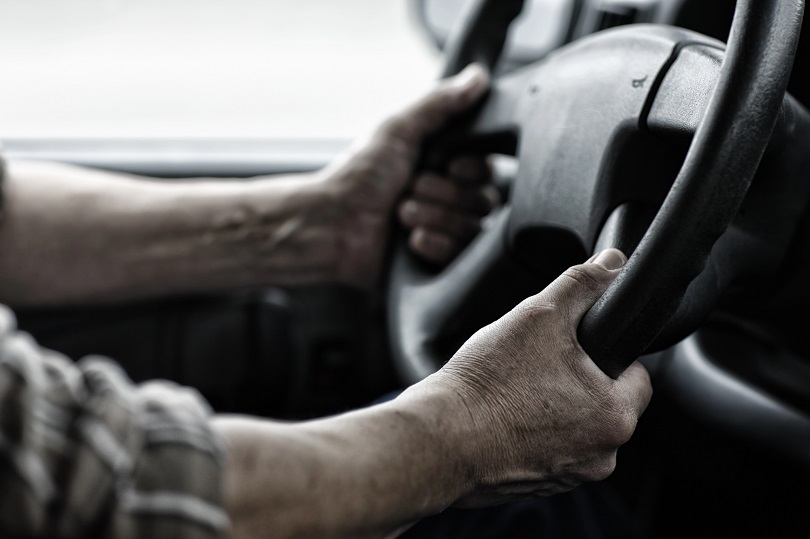Imagem das mãos de um homem posicionadas sobre o volante de um veículo. 