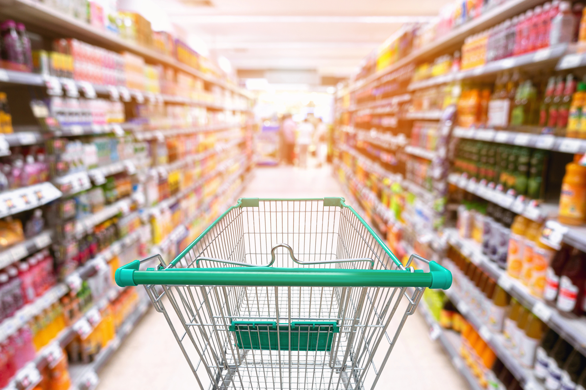 Carrinho de compras em primeiro plano em um corredor de supermercado com prateleiras repletas de produtos.