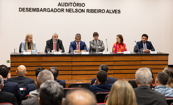 Plateia com os participantes. No fundo, a mesa com as autoridades, cor madeira, a parede é bege.