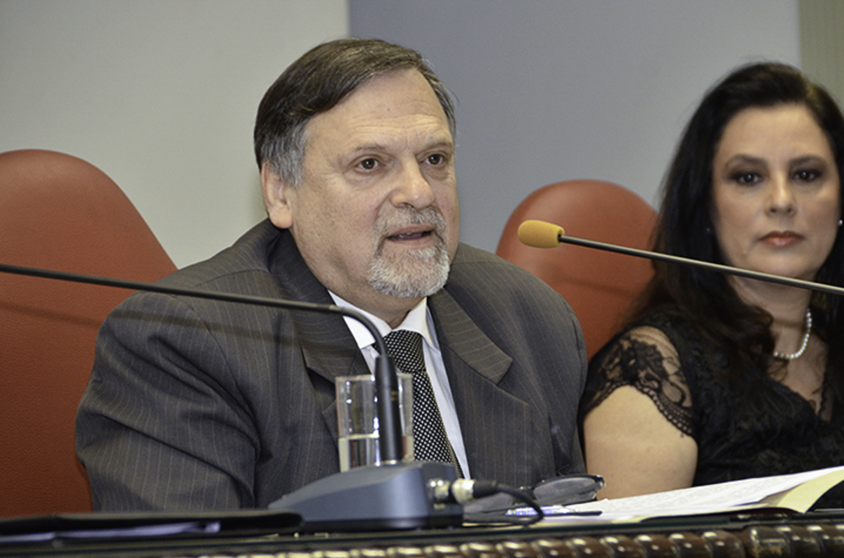 Edson Lerrer é branco, usa cabelo castanho curto liso, terno cinza, camisa branca e gravata xadrez. está falando ao microfone. A seu lado, em segundo plano, aparece a desembargadora Luciane Barzotto.  