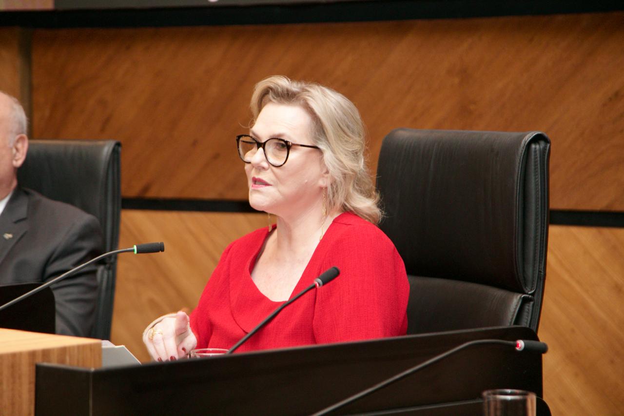 Laís Helena é branca, loira de cabelos lisos e usa óculos de armação redonda e preta. Está de de perfil e fala ao microfone.