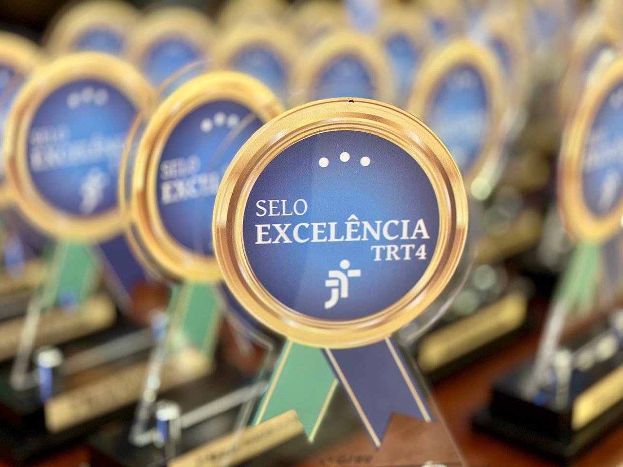 Diversos troféus sobre a mesa. Um deles está em foco. A arte é formada por um círculo dourado, com duas fitas cruzadas abaixo nas cores verde e azul. No centro está escrito Selo Excelência TRT4, com o símbolo da Justiça do Trabalho.