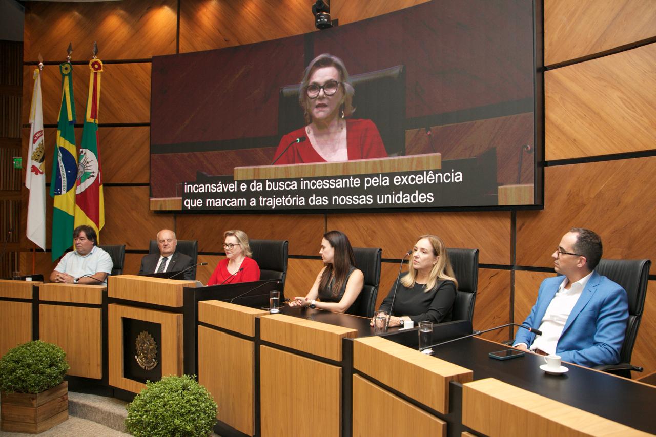 Seis pessoas aparecem na mesa oficial. No canto esquerdo estão as bandeiras de Poa, do Brasil e do RS.