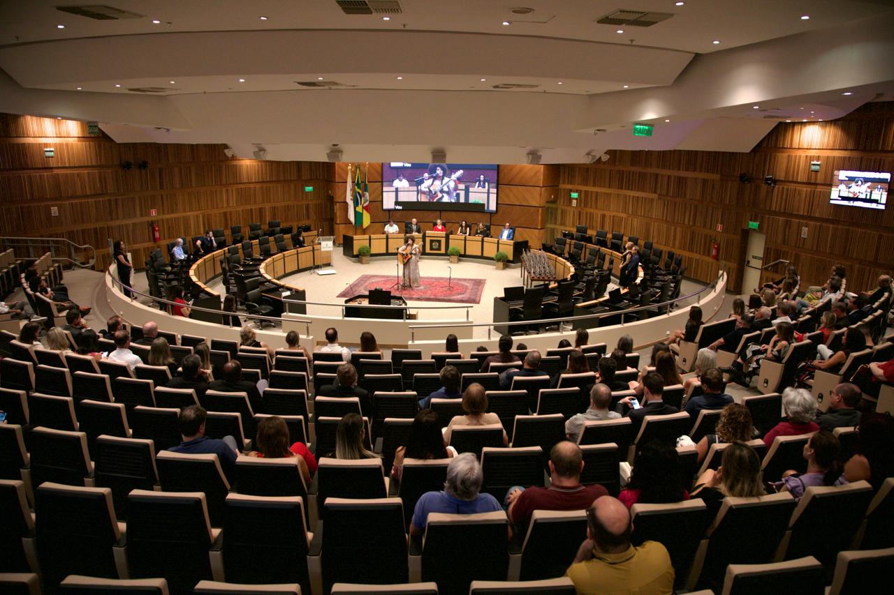 Foto geral do plenário. O público aparece de costas e, ao fundo, Chryssie está no meio da nave central cantando e tocando violão. Atrás dela aparece a mesa oficial e o telão.