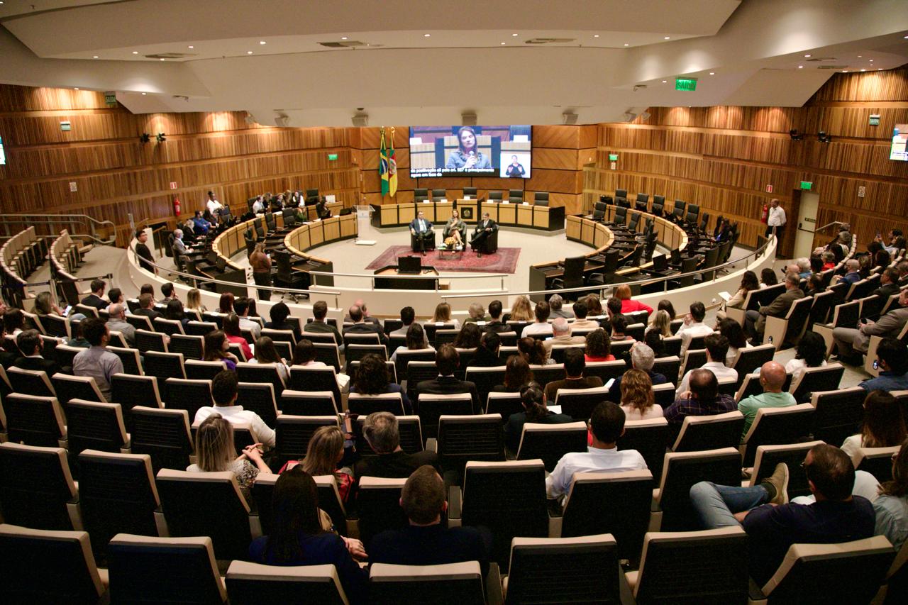 Foto geral do plenário, com público em primeiro plano e palestrantes ao fundo na nave central.