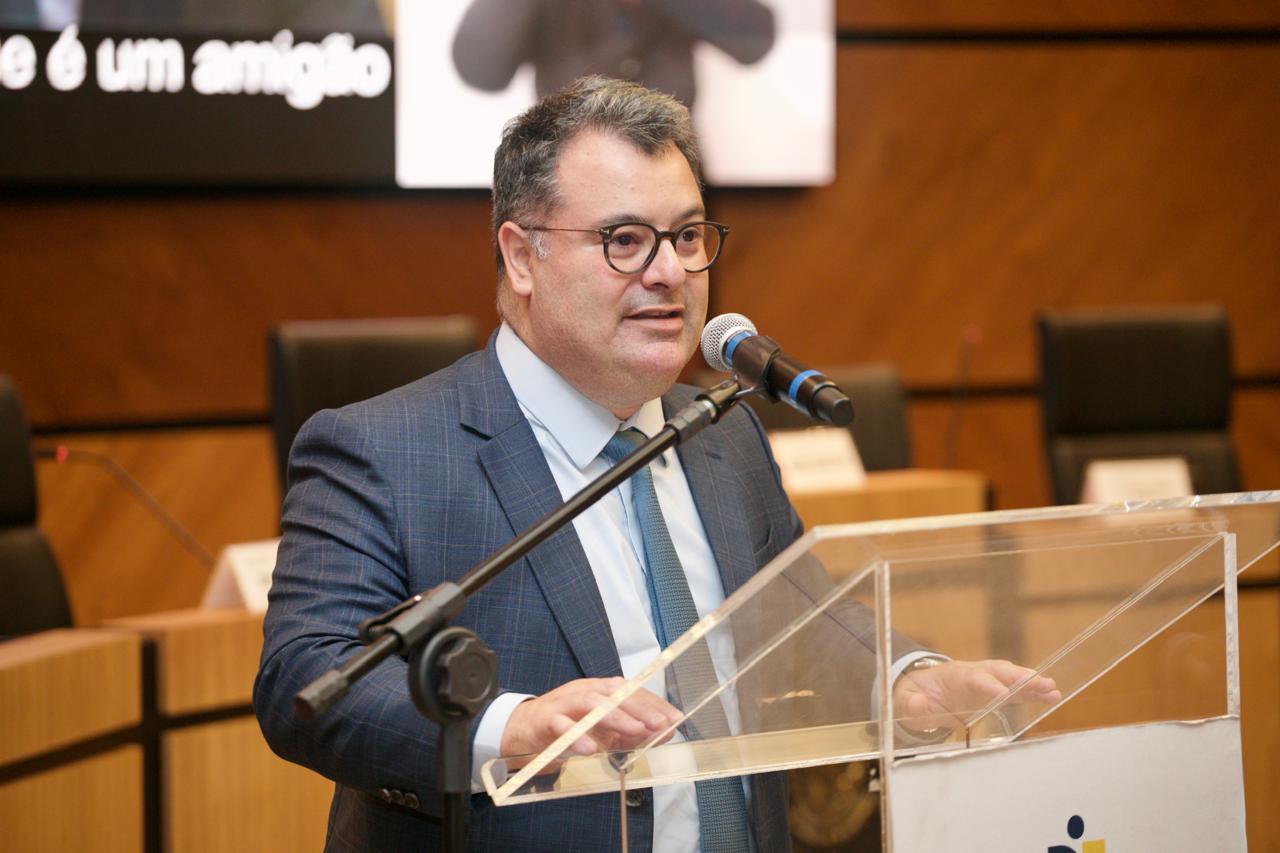 Foto de um homem branco de cabelo grisalho. Ele usa blazer azul xadrez, camisa social branca, e gravata azul clara. Está com as mãos repousadas sobre uma bancada de acrílico. Ao fundo aparecem poltronas, bancadas de madeira, o painel parede de madeira do plenário e um pedaço do telão.