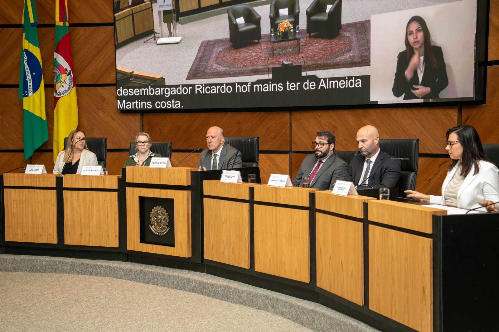 Os seis magistrados aparecem sentados atrás da bancada de madeira. Ao centro, o presidente Martins Costa fala ao microfone. Na esquerda aparecem as bandeiras do Brasil e do RS, e atrás dos magistrados aparece um telão ligado.