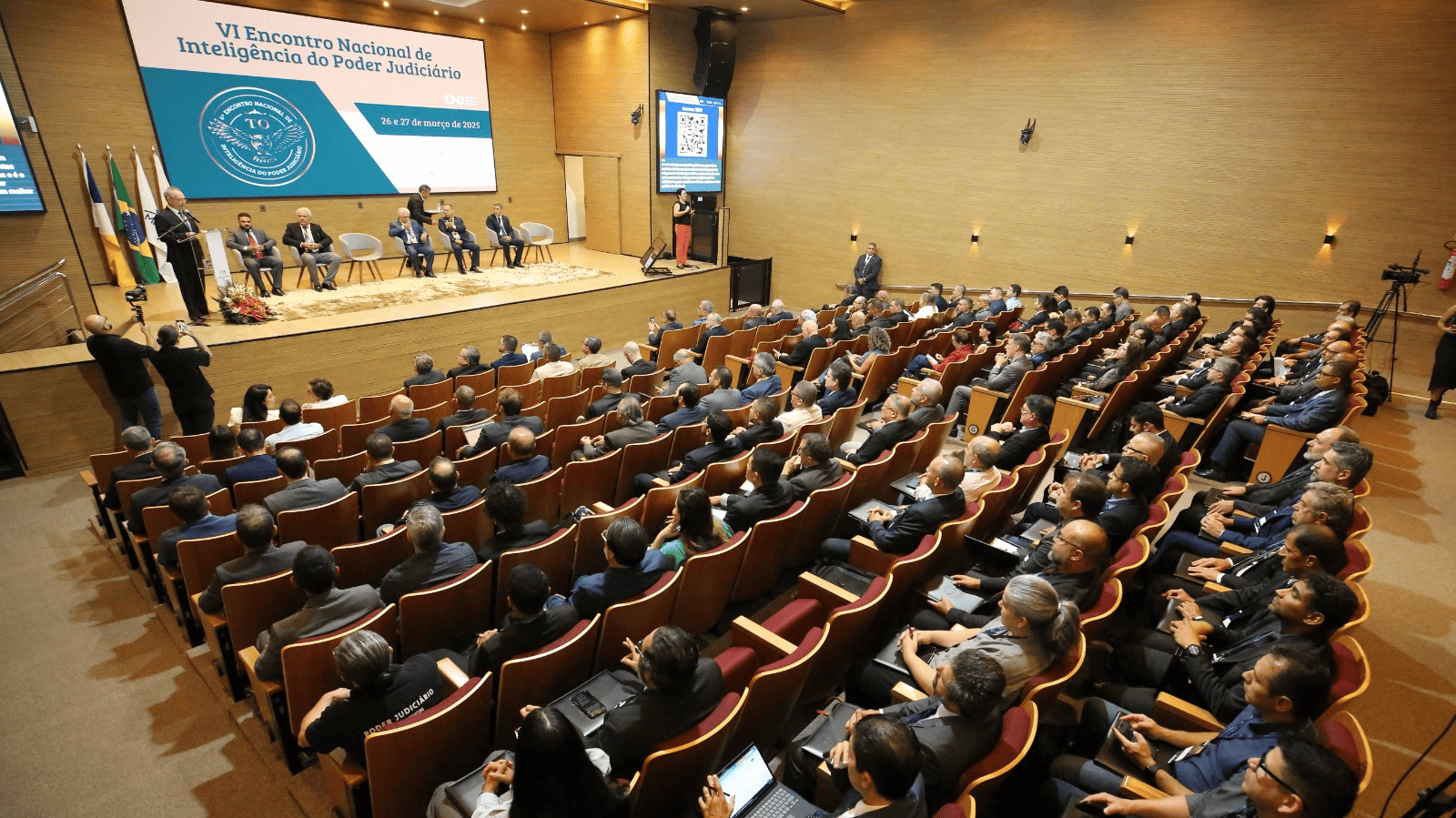 Auditório lotado. Ao fundo, o banner escrito "Inteligência e Segurança no Judiciário". No palco também existem bandeiras não hasteadas.