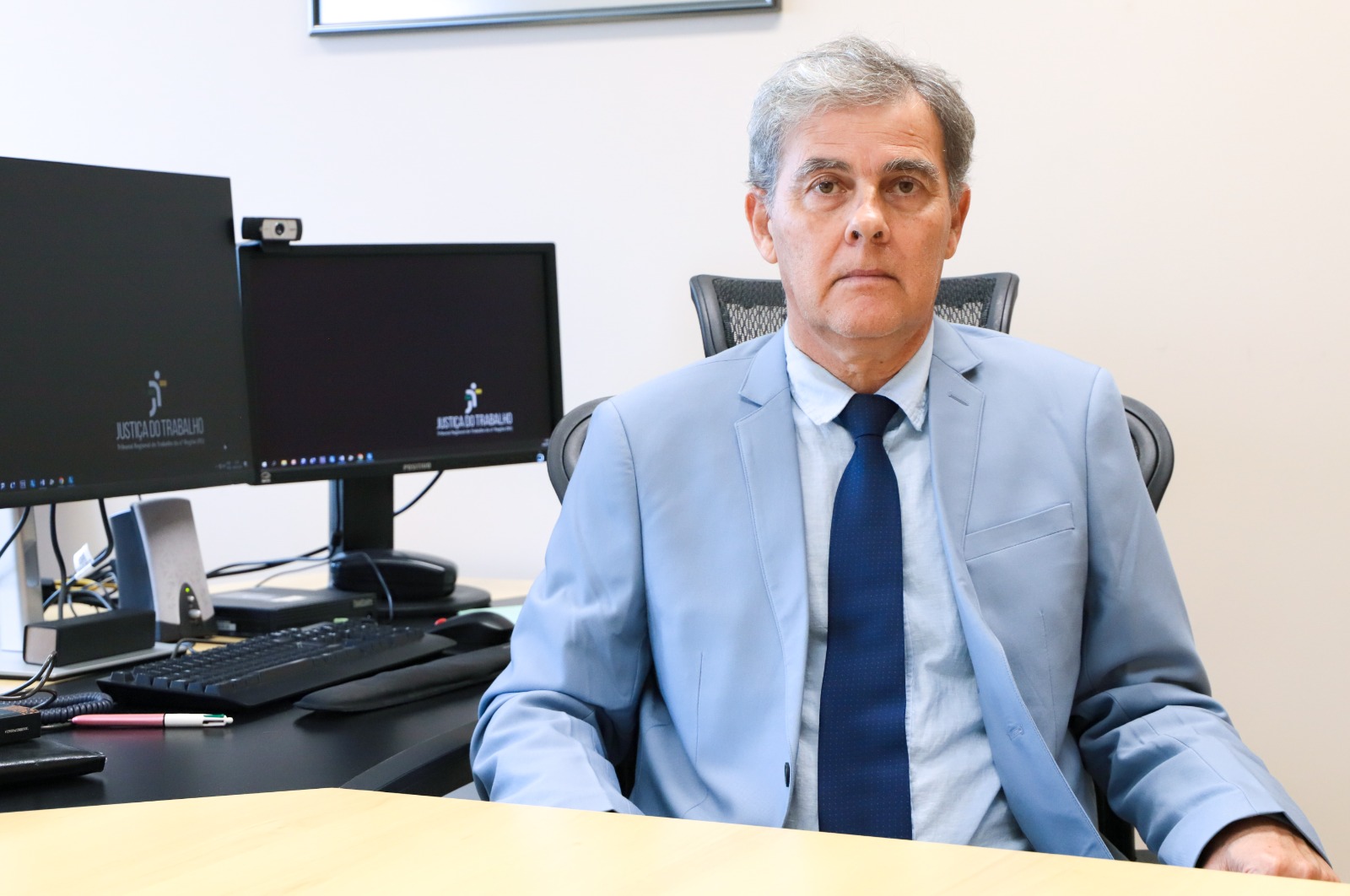 A imagem mostra o desembargador Ricardo Carvalho Fraga sentado em seu gabinete. Ele veste um terno azul claro, camisa azul e uma gravata azul escura. Ele tem uma expressão séria e olha diretamente para a câmera. No fundo, há uma mesa com um teclado, um mouse, uma caneta e dois monitores ligados, ambos exibindo o logotipo da Justiça do Trabalho. A cadeira em que ele está sentado tem um encosto de malha.