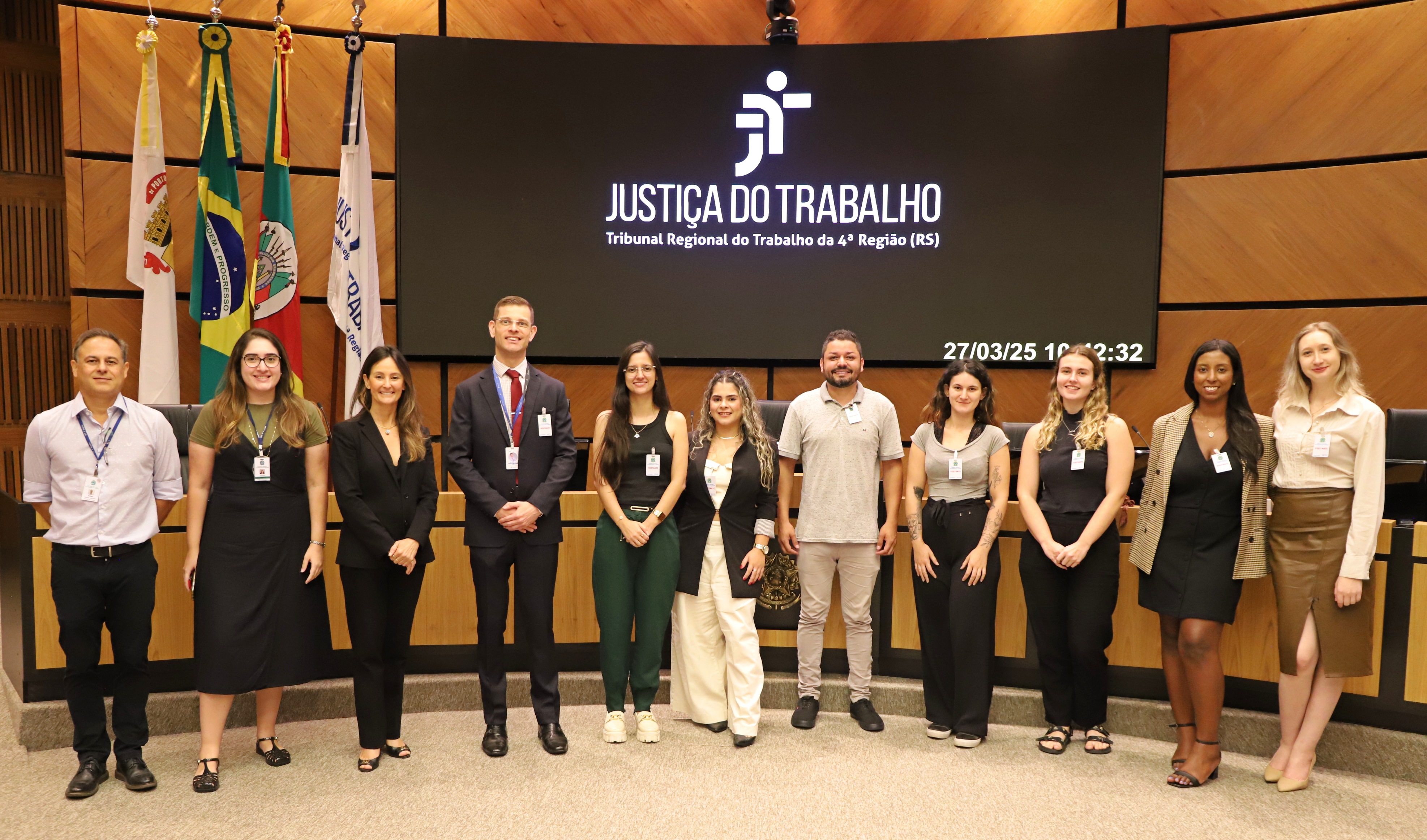 Equipe do Cerimonial, universitários, professor e juíza em visita ao palco do plenário do Tribunal Regional do Trabalho da 4ª Região, atrás deles, um telão com a logo da Justiça do Trabalho do RS.