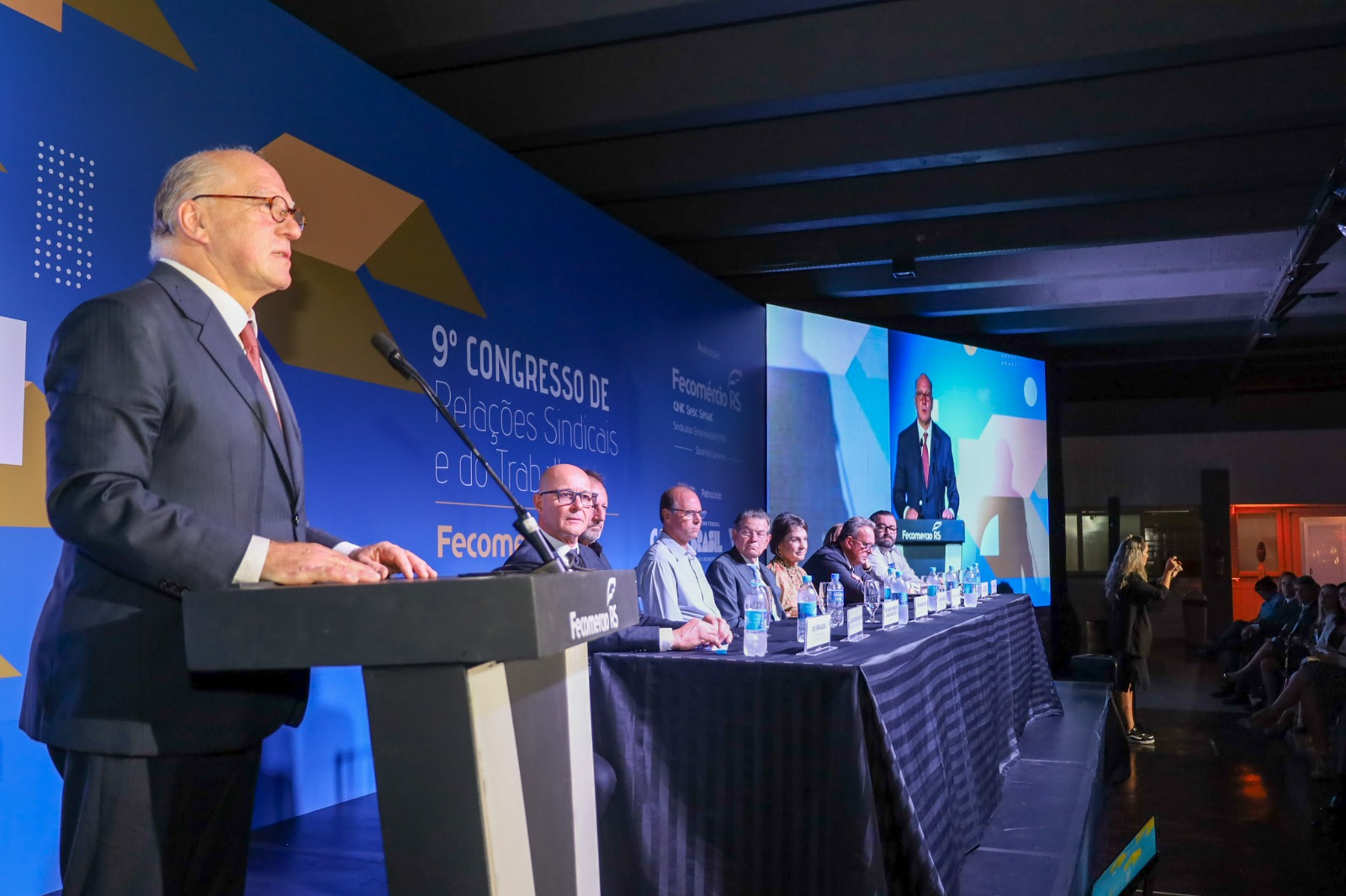 Presidente discursa no púlpito com a mesa de autoridades de fundo