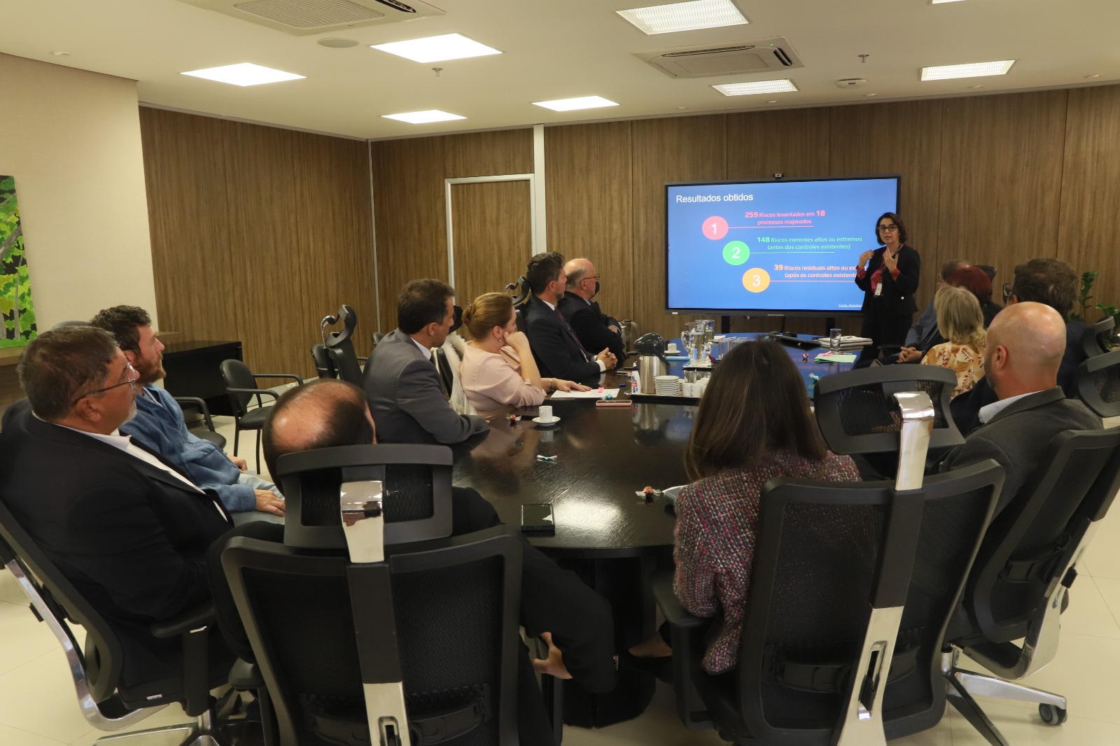 A servidora Tatiana Patricia Krause faz apresentação, com telão de fundo, aos presentes na reunião, no Salão Nobre do TRT-4. Ela estás de pé e os demais sentados ao redor de uma mesa.