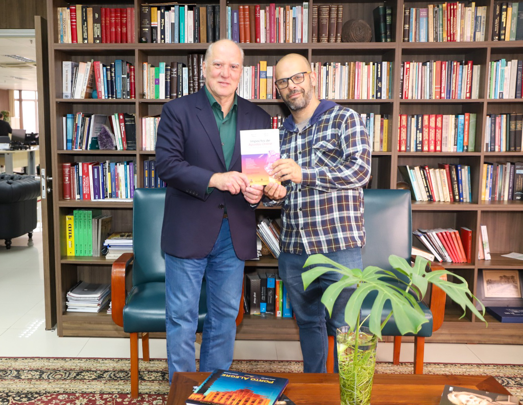 O desembargador Ricardo Martins Costa (à esquerda) e o juiz do Trabalho Oscar Krost (à direita) posam sorridentes durante a entrega do livro Impactos das Apostas Online no Direito do Trabalho, no gabinete da Presidência do TRT-RS.